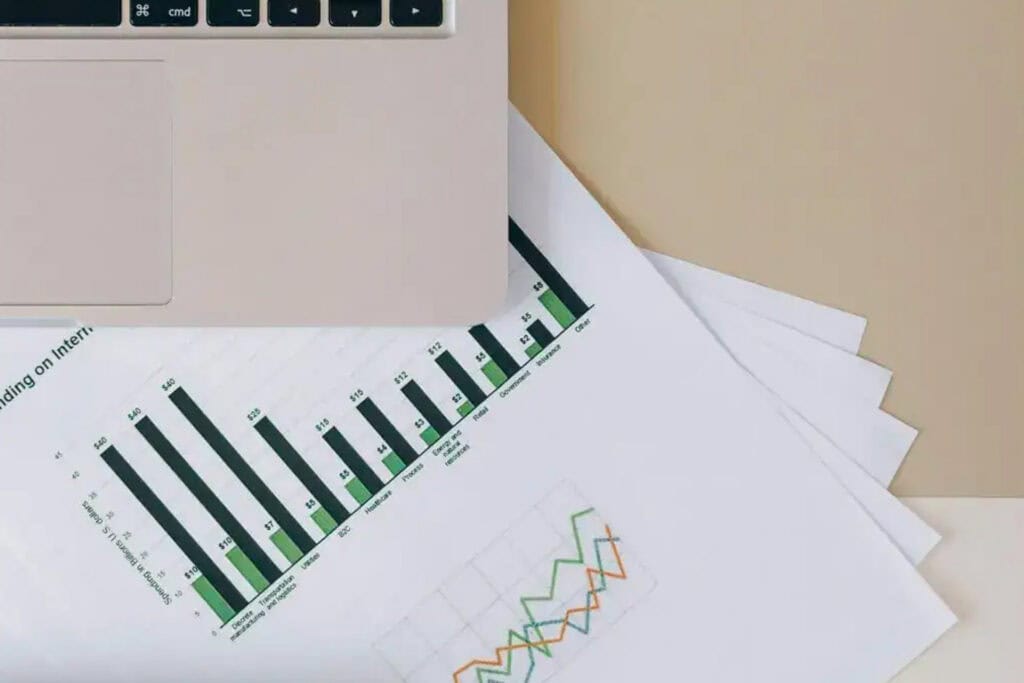 Laptop keyboard and trackpad partially visible above printed business reports showing bar and line charts with green and dark-colored data visualizations on a desk.